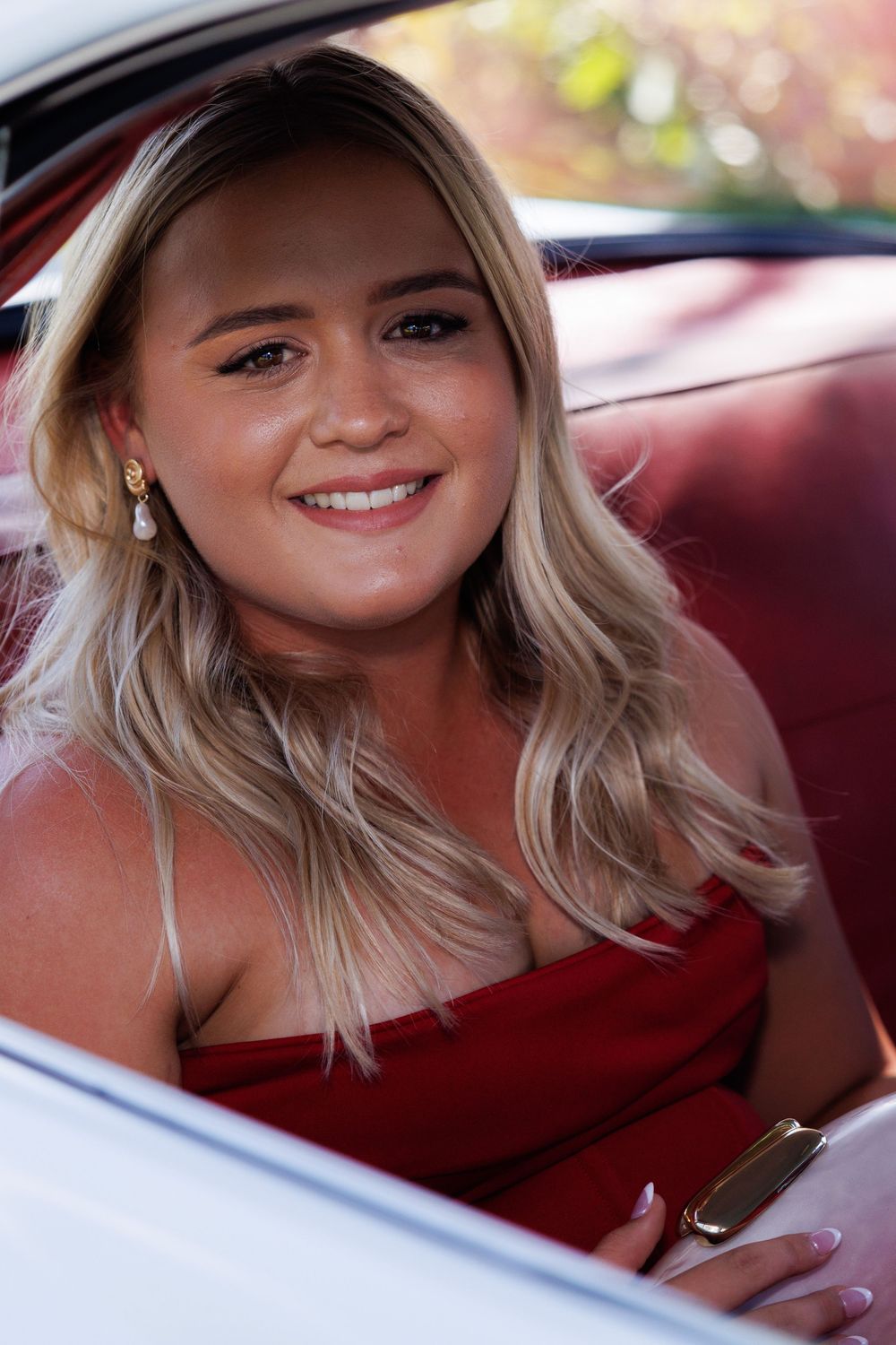 Smiling young woman with long blonde hair in a red strapless dress sits in a vintage car with red upholstery.