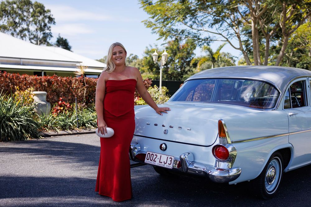 Young woman in a long red formal dress stands beside a vintage cream car with a licence plate on a sunny driveway.
