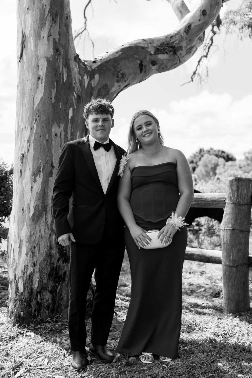 Black and white portrait of a young couple in formal wear posing beside a large eucalyptus tree and wooden fence.