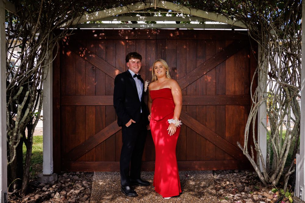 Young couple in a black tuxedo and red evening gown pose under a vine-covered arbor in front of rustic wooden barn doors.