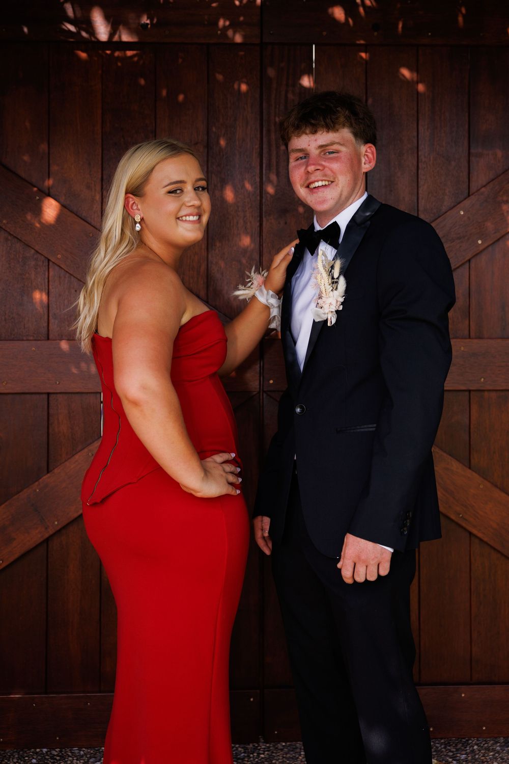 Smiling young man in a tuxedo with a floral boutonniere stands beside a woman in a red gown against a wooden barn door.
