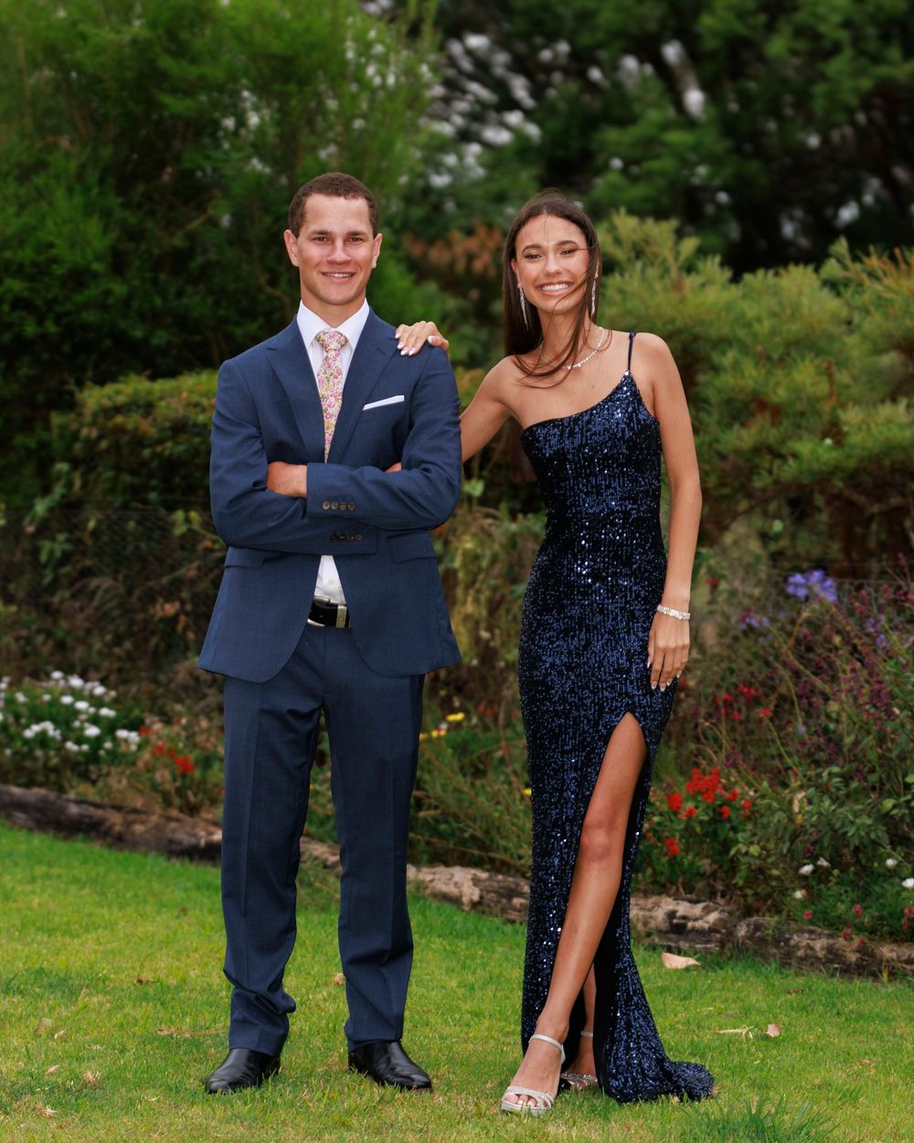 Couple poses on a garden lawn, a young man in a navy suit beside a woman in a navy sequin gown with a high slit.