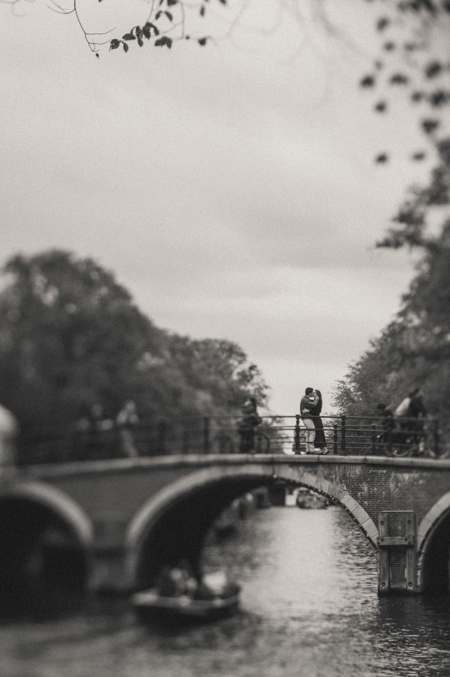 Black and white photo of a couple embracing on an arched canal bridge in Amsterdam, with a small boat on the water below.