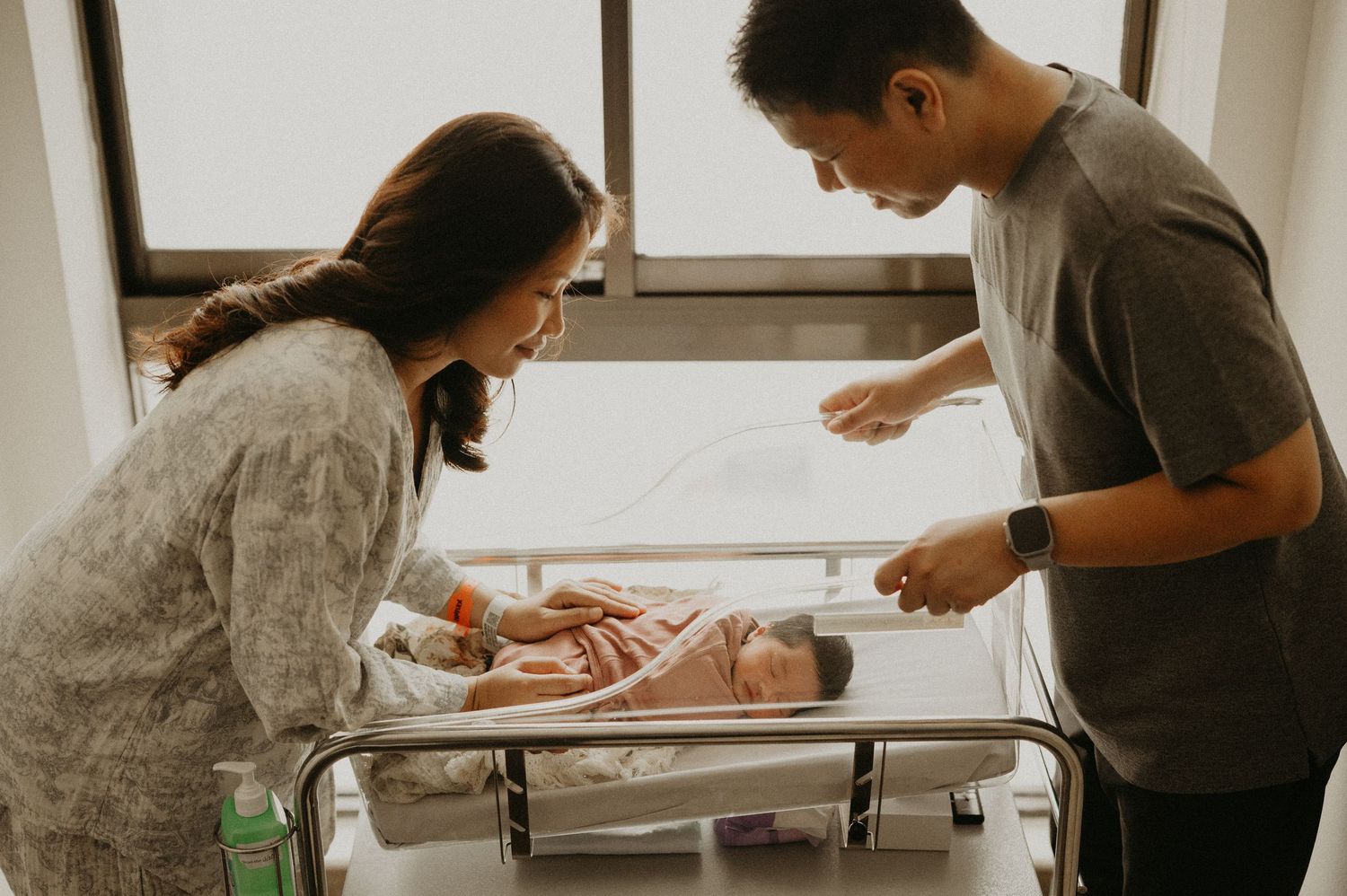 Smiling parents gaze lovingly at their sleeping newborn baby wrapped in a blanket in a hospital bassinet beside a bright window.