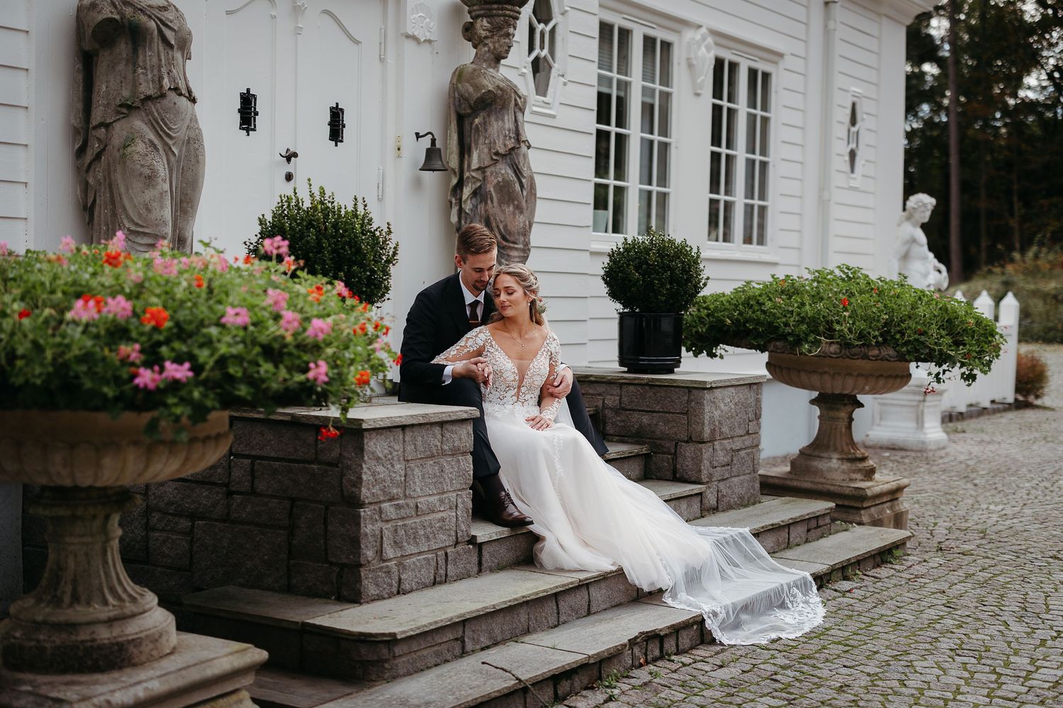 Bride in a lace gown and groom in a dark suit sit together on stone steps outside a white manor with flower-filled urns.