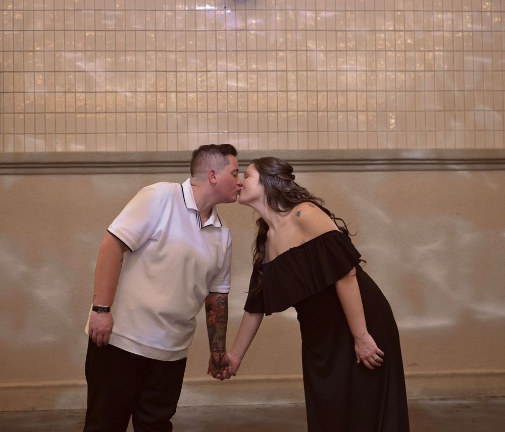 A couple sharing a romantic kiss while holding hands in front of a tiled wall in an urban subway setting.