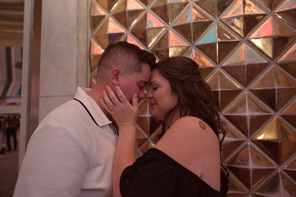 Romantic couple sharing an intimate moment, foreheads touching, posed against a shimmering geometric gold mosaic wall.