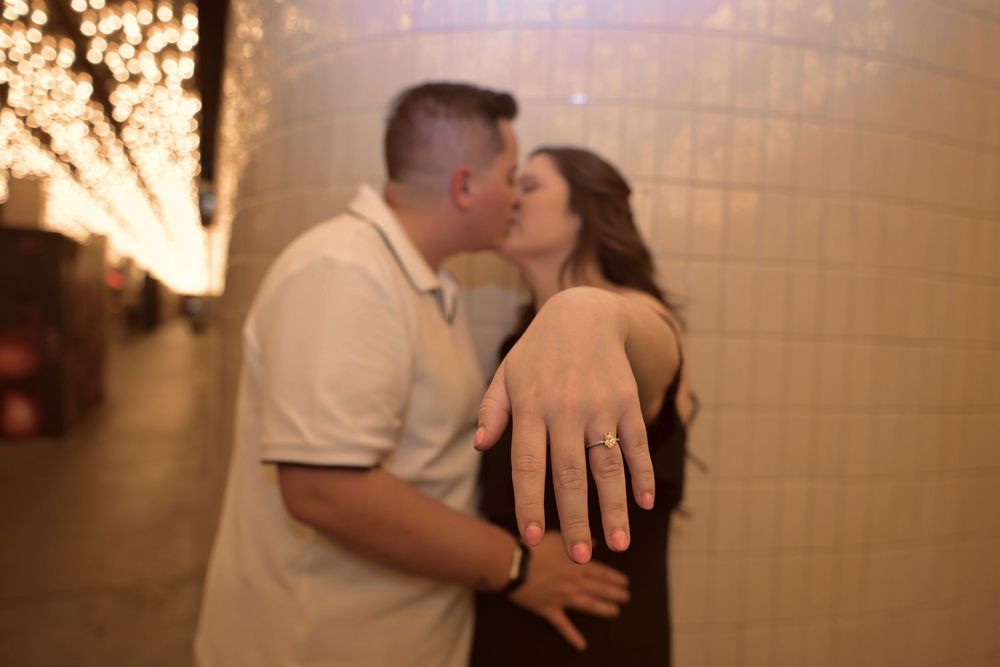 Couple sharing a romantic kiss during engagement proposal, woman displaying diamond ring on her hand in a warmly lit tunnel.