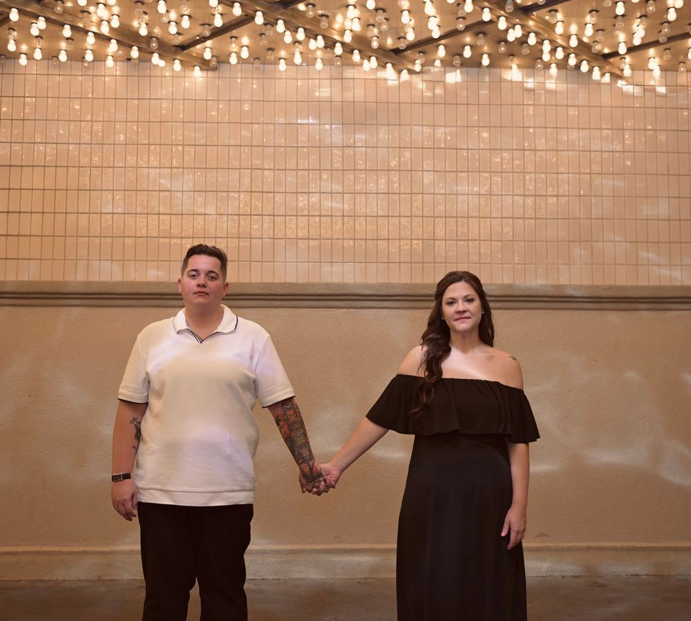 Couple holding hands standing against a tiled wall with string lights, man in white polo and woman in black off-shoulder dress.