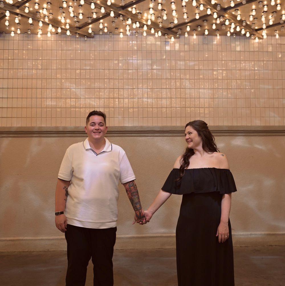 Couple holding hands under string lights in front of a tiled wall.