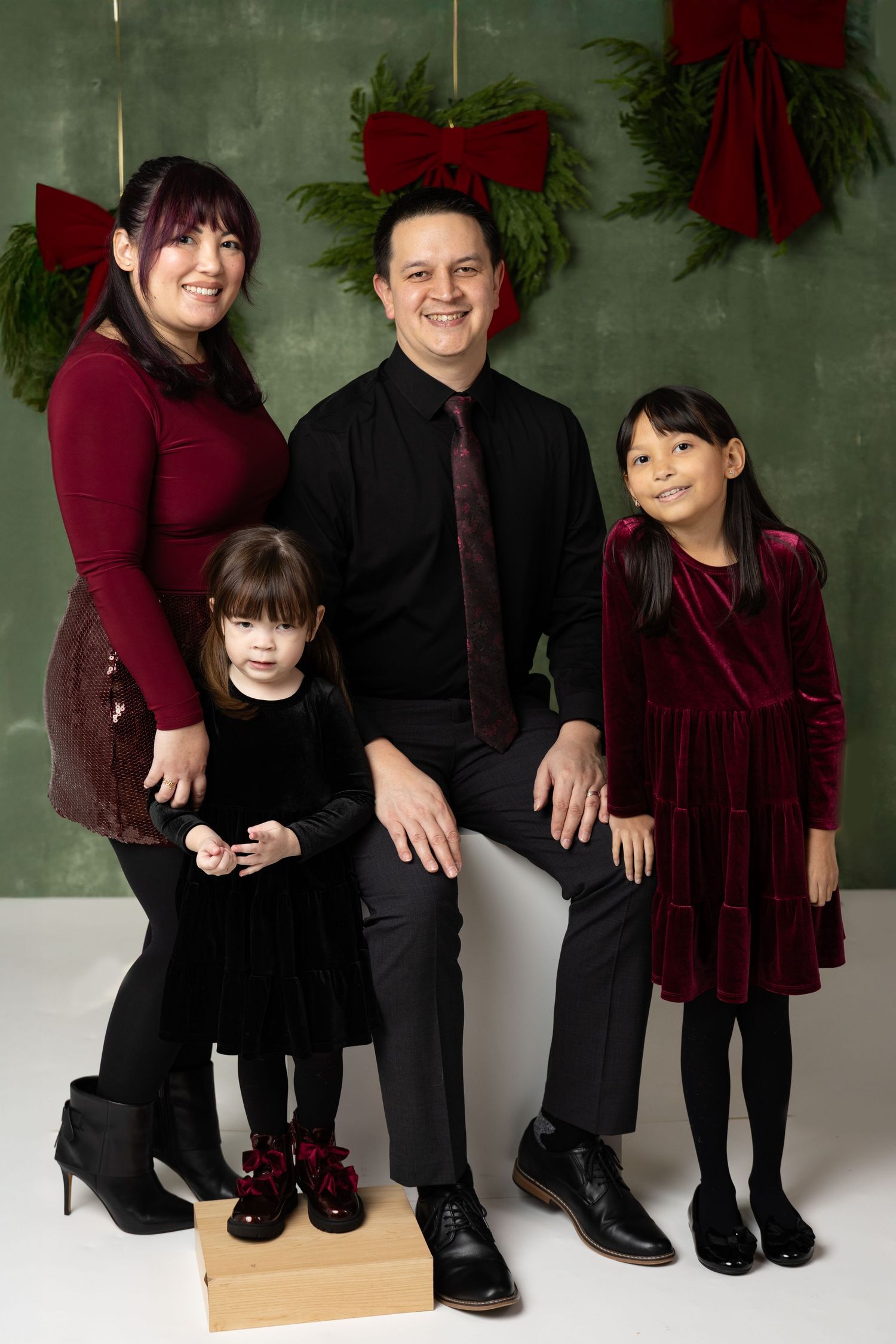 Family of four posing for a Christmas portrait in coordinating burgundy and black outfits with holiday greenery backdrop.