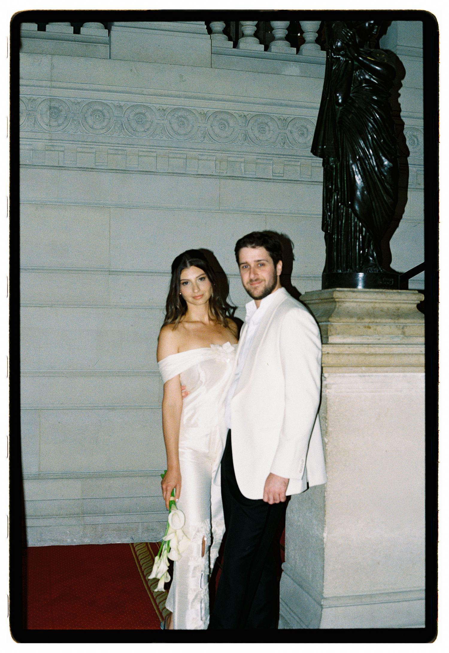 Elegant couple posing on grand staircase, woman in white off-shoulder gown holding flowers, man in white tuxedo jacket.