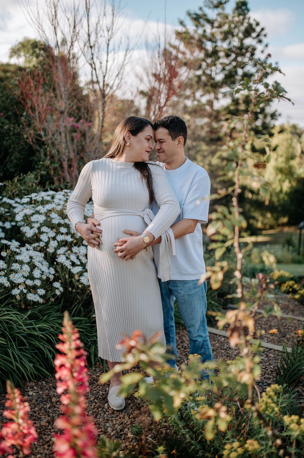 Couple sharing a tender moment in a garden, with the woman in a white ribbed dress cradling her baby bump.