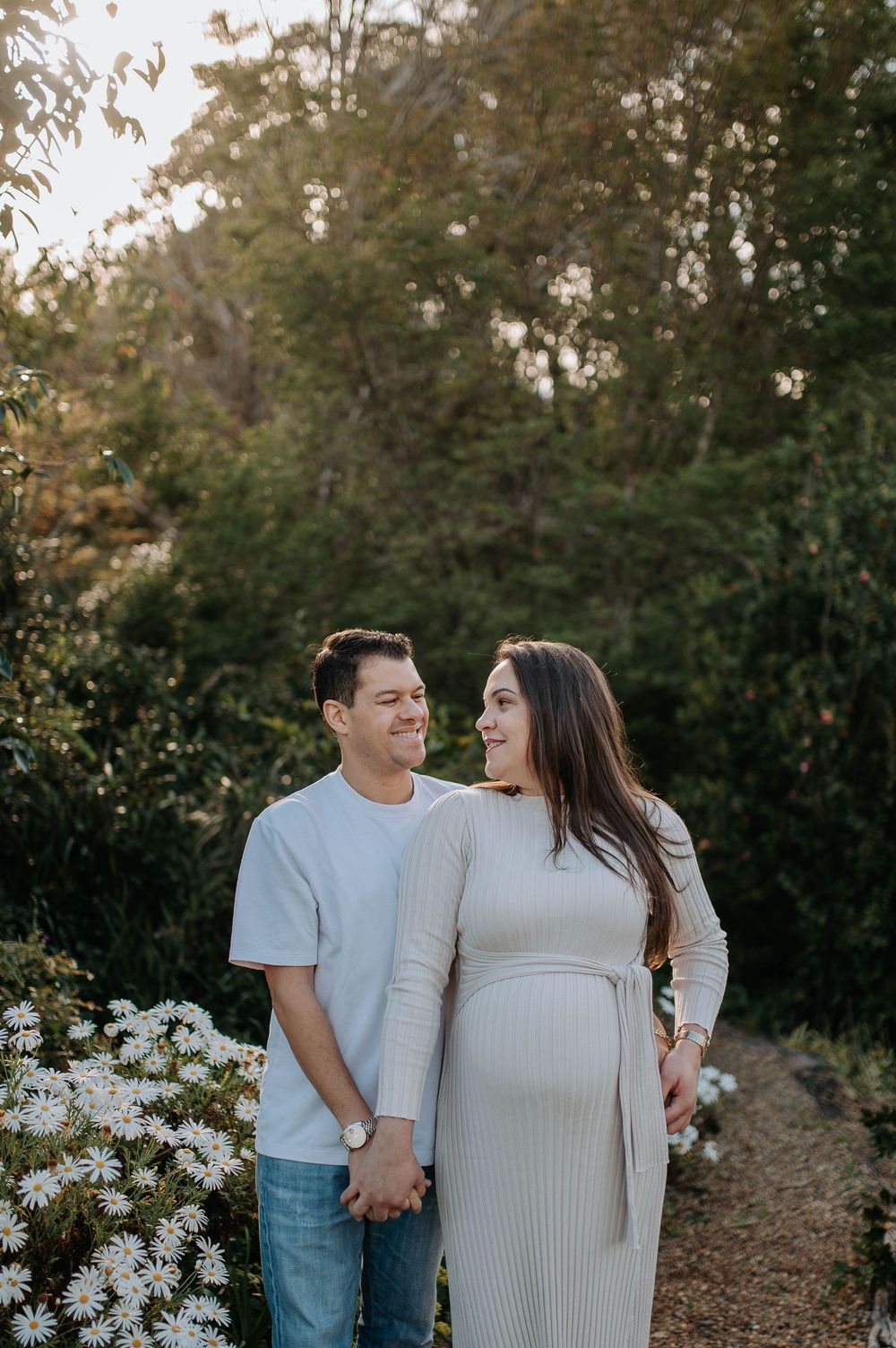 Couple holding hands in a garden path, woman in cream ribbed maternity dress, man in light blue tee and jeans, surrounded by greenery.