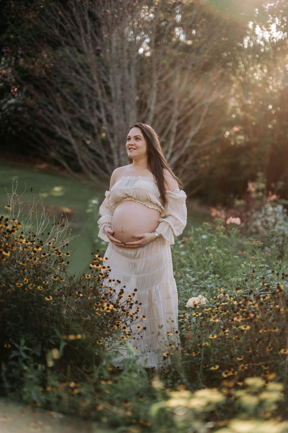 Pregnant woman in white dress holding baby bump in sunlit garden with wildflowers and lush greenery.