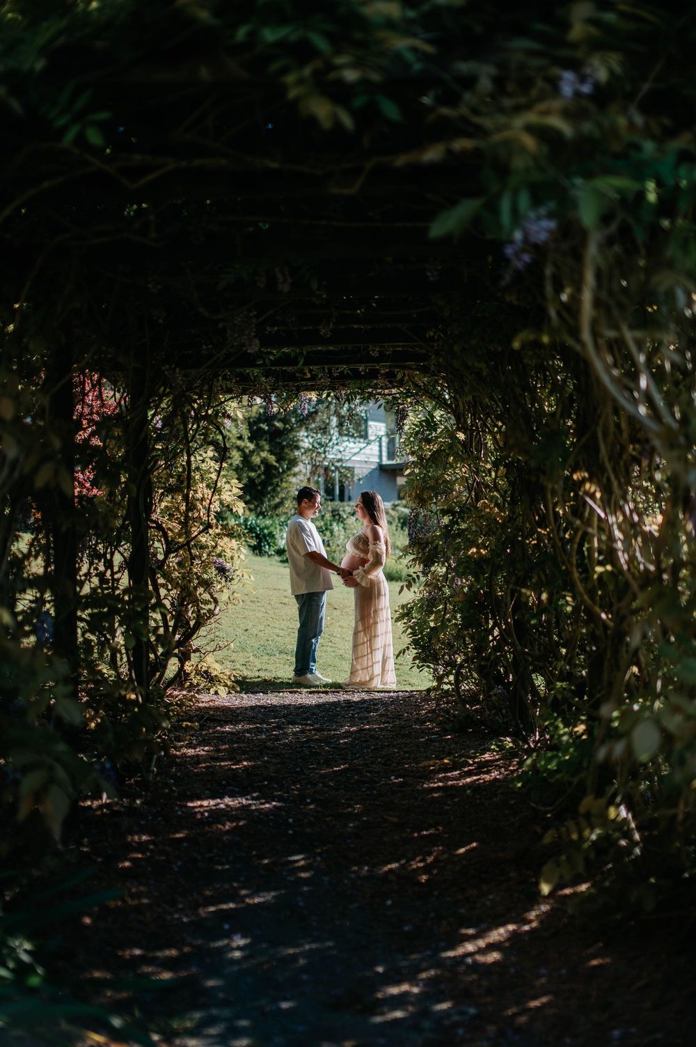 Couple holding hands framed by a lush green garden tunnel archway with sunlight illuminating them from behind.