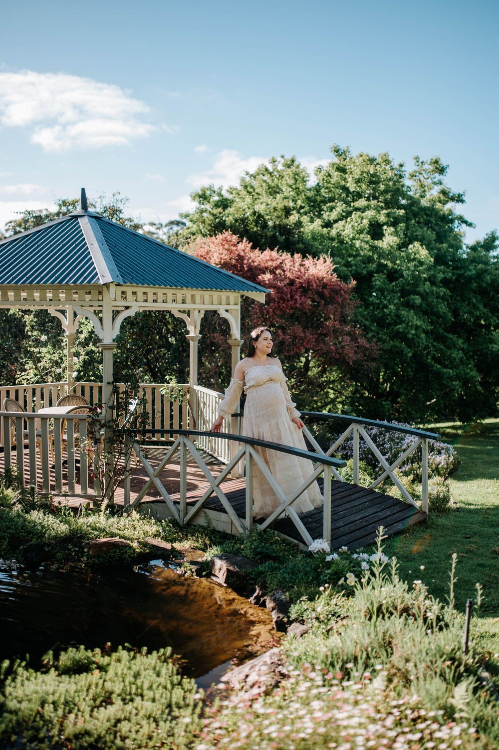 Pregnant woman in elegant gown standing on garden bridge near white gazebo surrounded by lush greenery on sunny day.