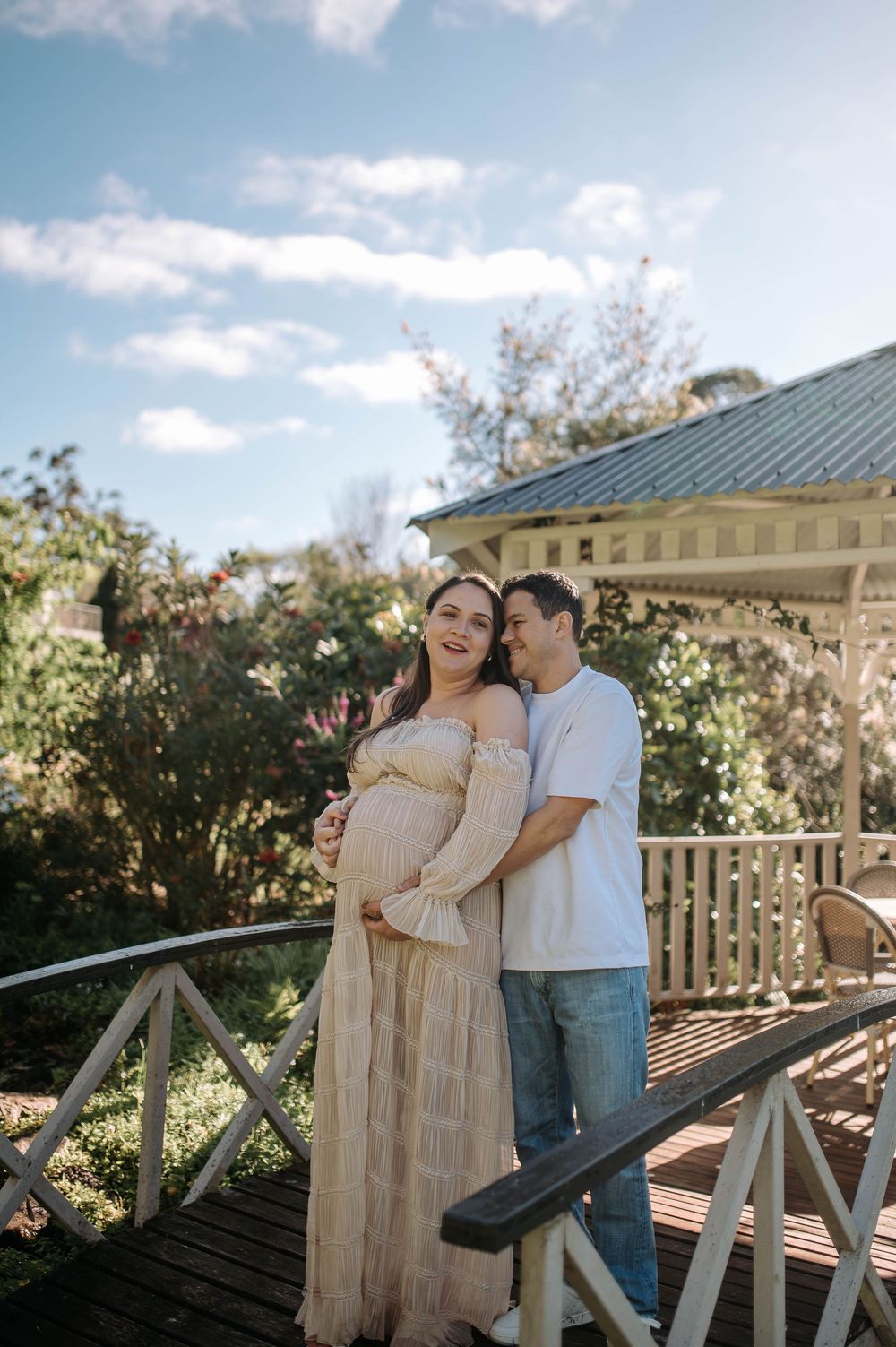 Couple poses on wooden bridge near gazebo, pregnant woman in flowy beige dress, man in white tee and jeans, lush garden backdrop.