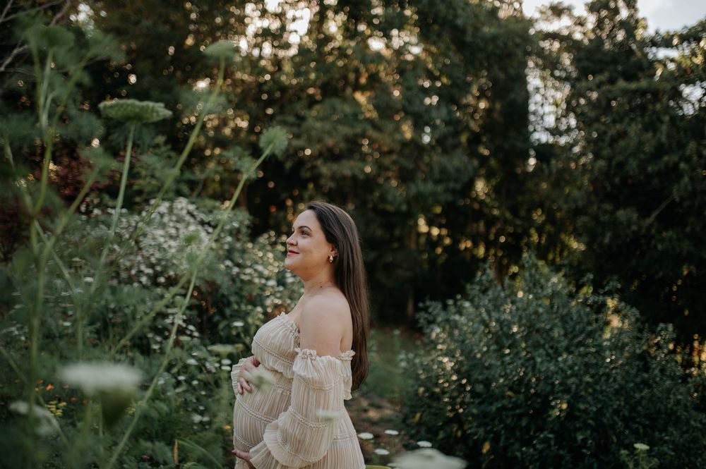 Pregnant woman in a flowy off-shoulder dress standing in a lush garden during golden hour.