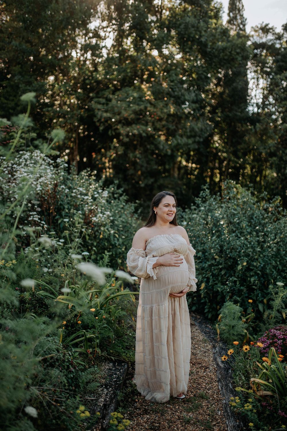 Pregnant woman in a flowing beige off-shoulder gown cradling her belly in a lush garden at golden hour.