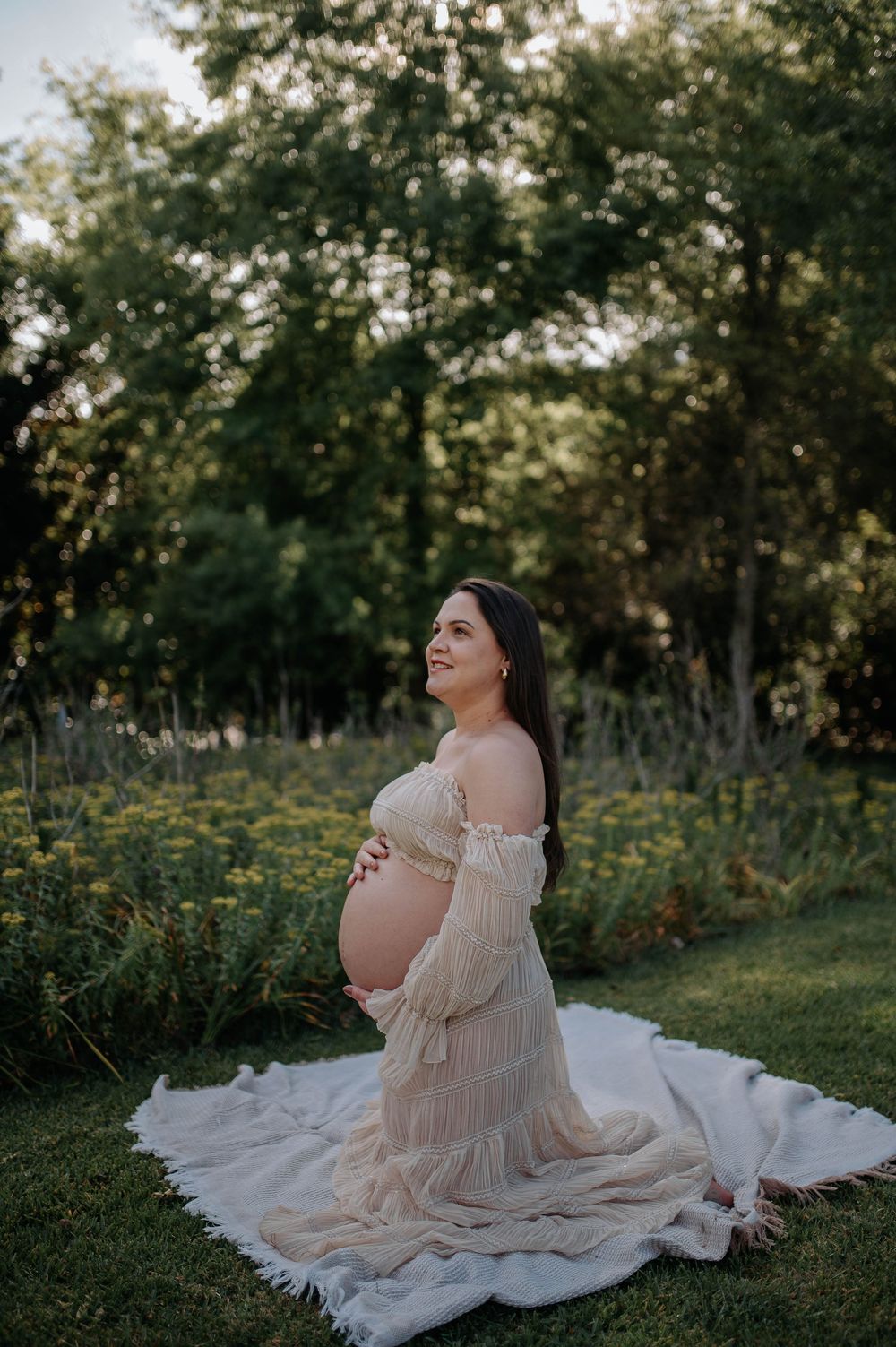 Pregnant woman in a flowing beige off-shoulder gown kneeling in a lush garden, cradling her baby bump.