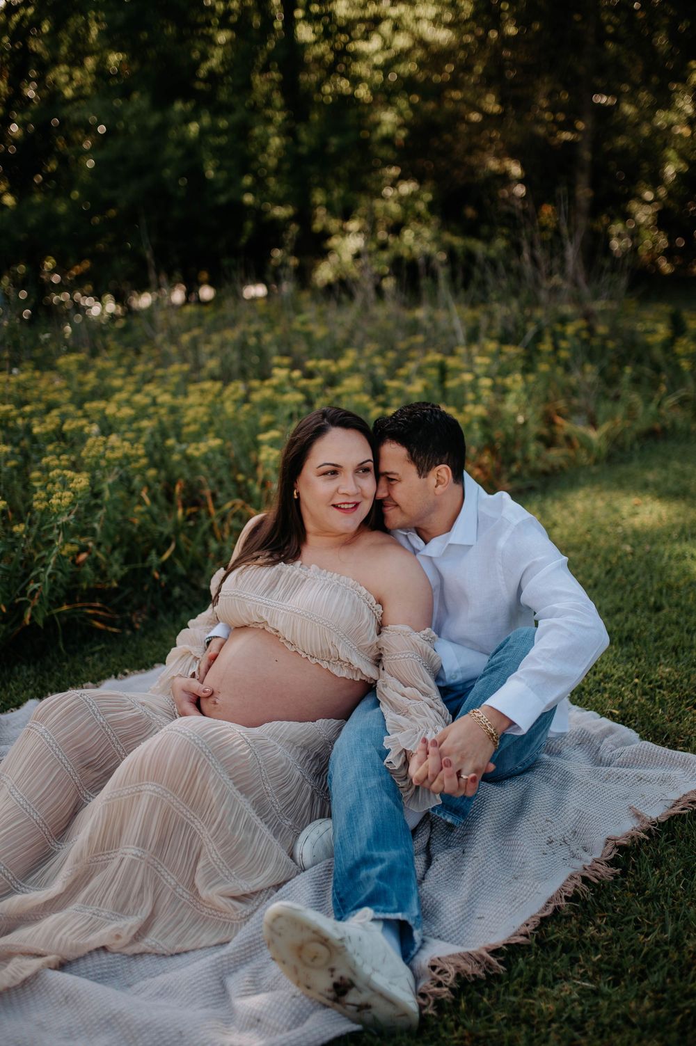 Pregnant couple sitting on blanket in garden, woman in sheer dress cradling baby bump while partner kisses her cheek.