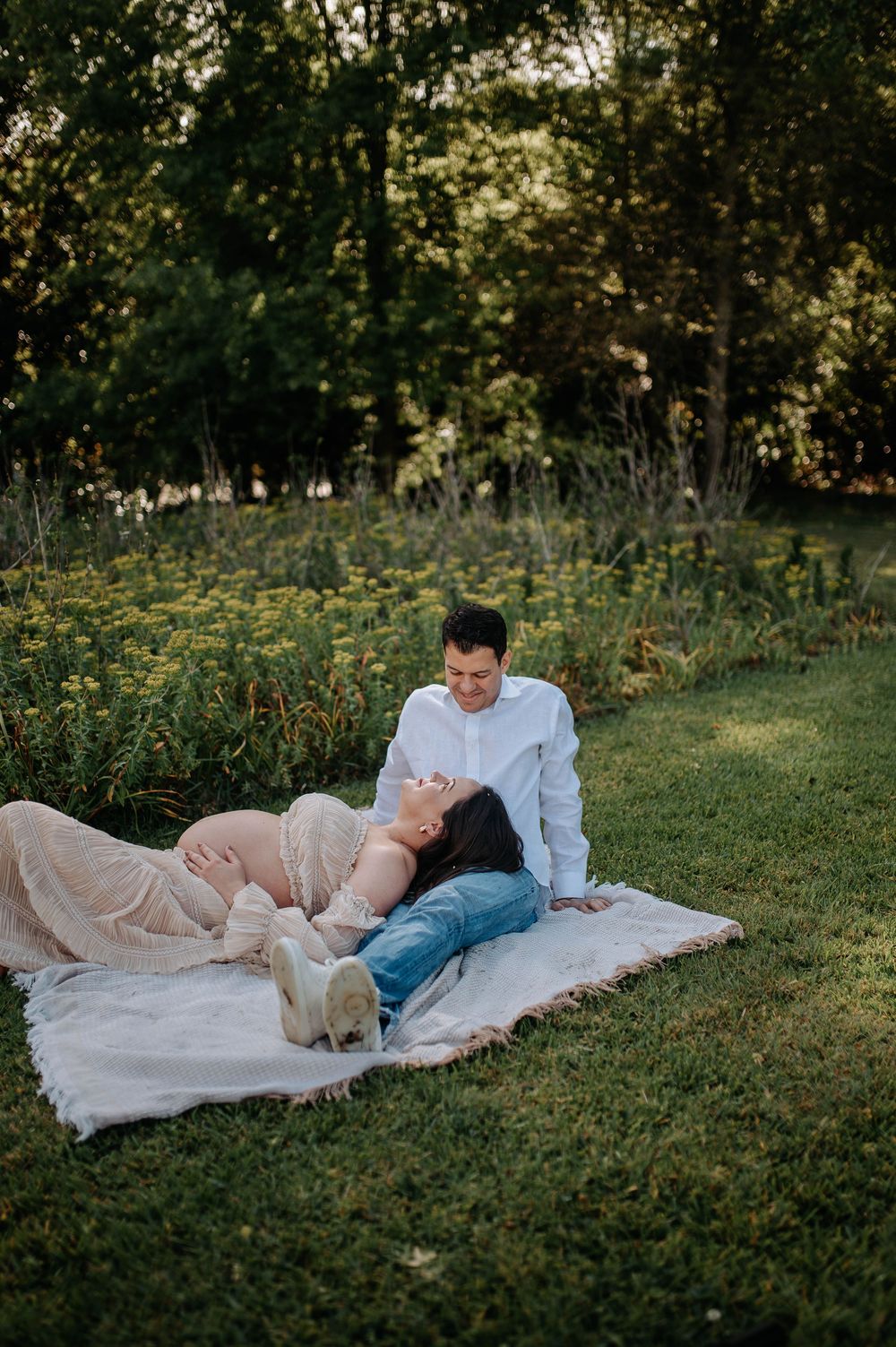 A pregnant woman lies on a blanket in a garden while her partner sits beside her, sharing a tender moment outdoors.