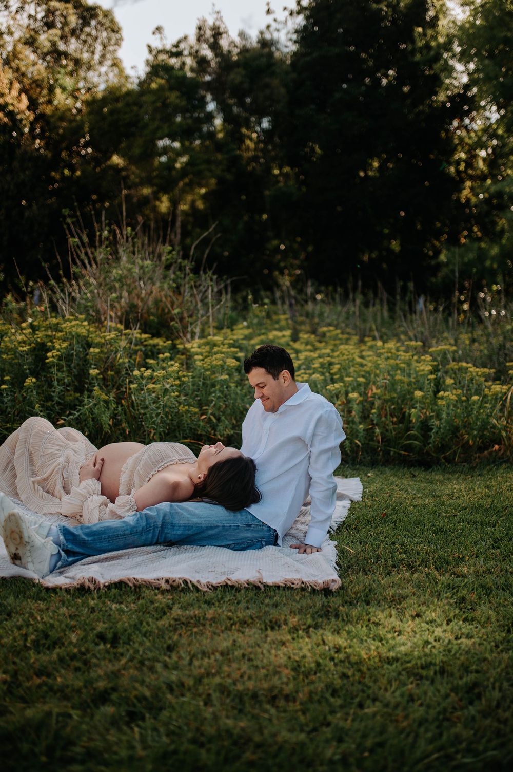 A pregnant woman lies on a blanket in a garden while her partner sits beside her, smiling warmly during a maternity photoshoot.
