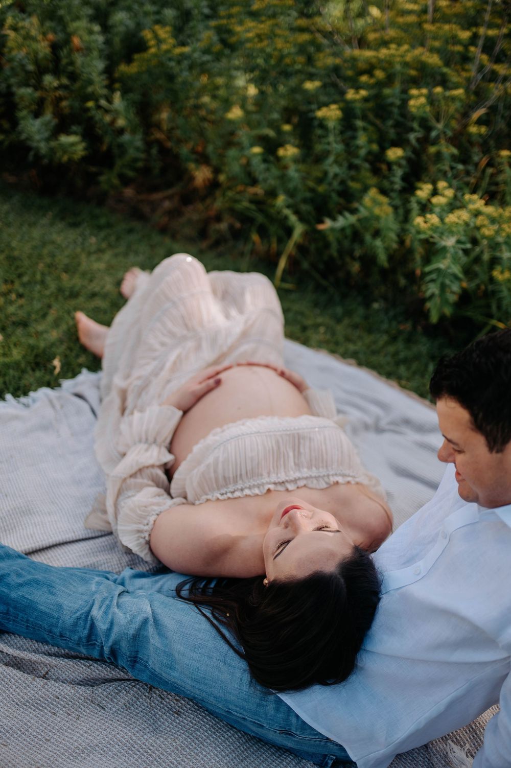 Pregnant woman in white dress lying on blanket outdoors, partner beside her, surrounded by lush green garden.