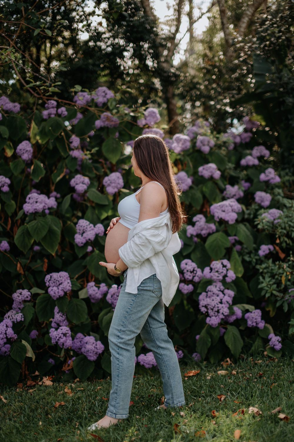 Pregnant woman in white crop top and jeans standing in front of purple hydrangea bushes outdoors.