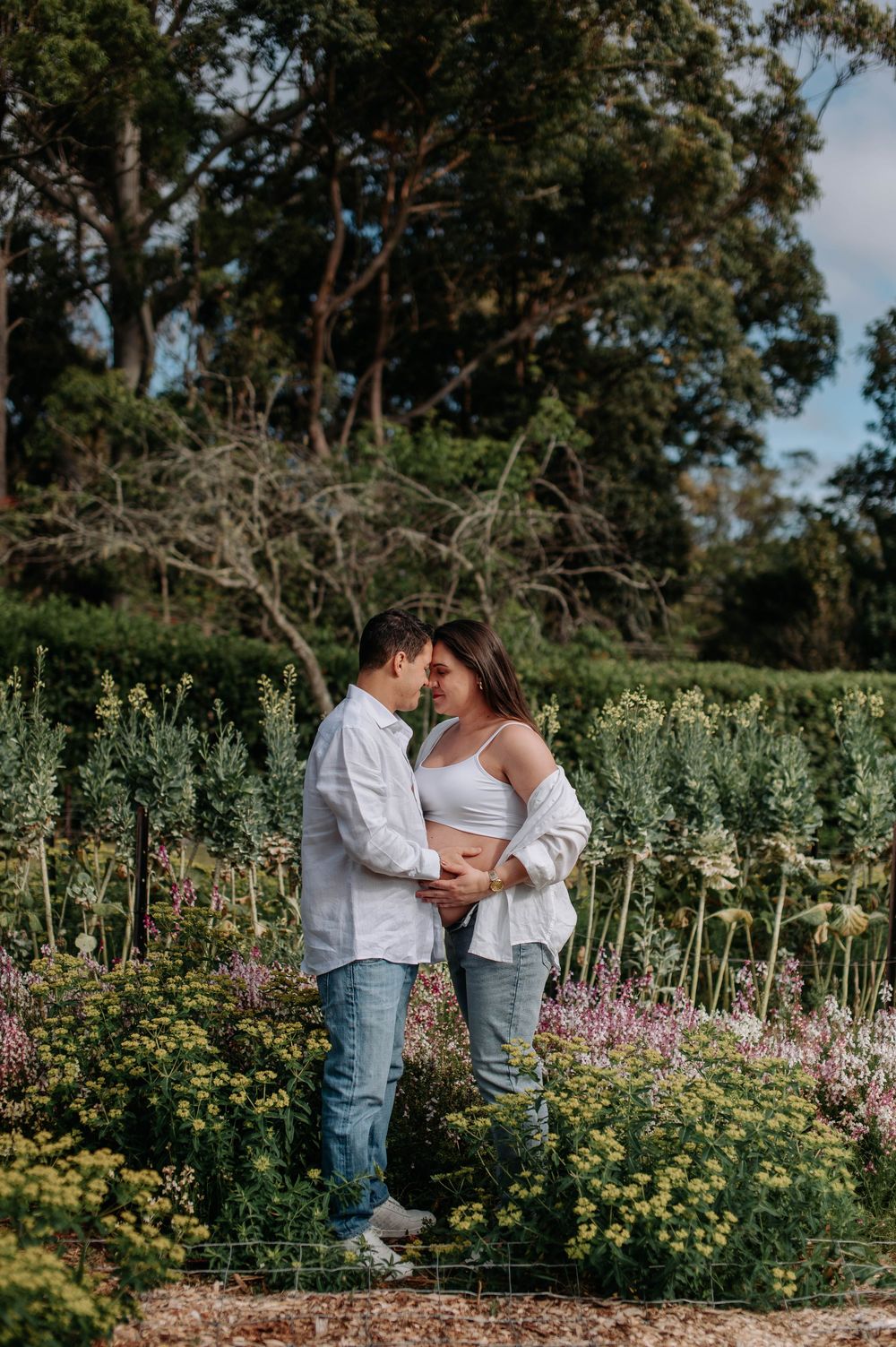 Couple in white shirts and jeans sharing a romantic moment in a lush garden with trees in the background.
