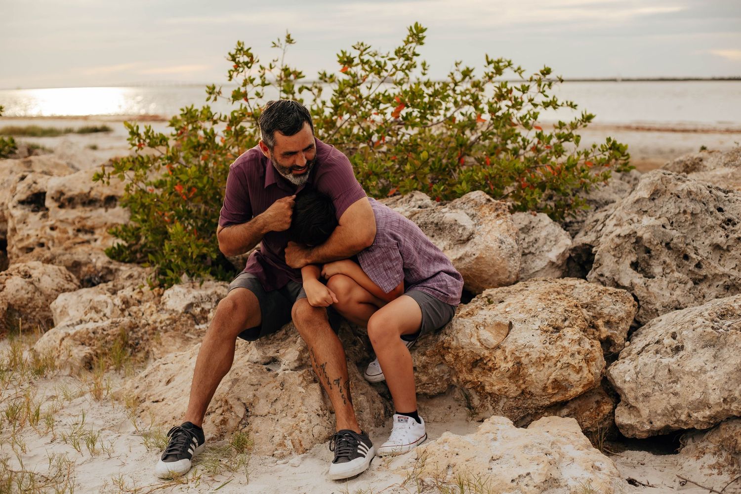 Two men share an affectionate embrace while sitting on coastal rocks near the water at sunset, surrounded by green shrubs and sandy ground.