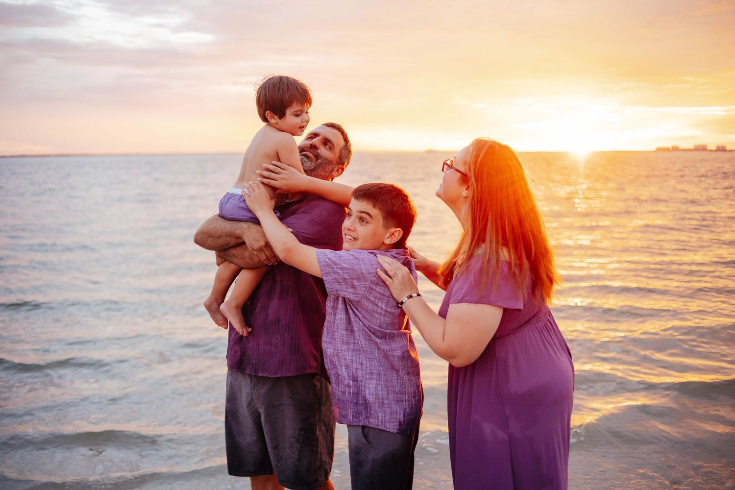 Black and white photo of a smiling family of four, with a toddler held in her mother's lap, posing close together on a beach.