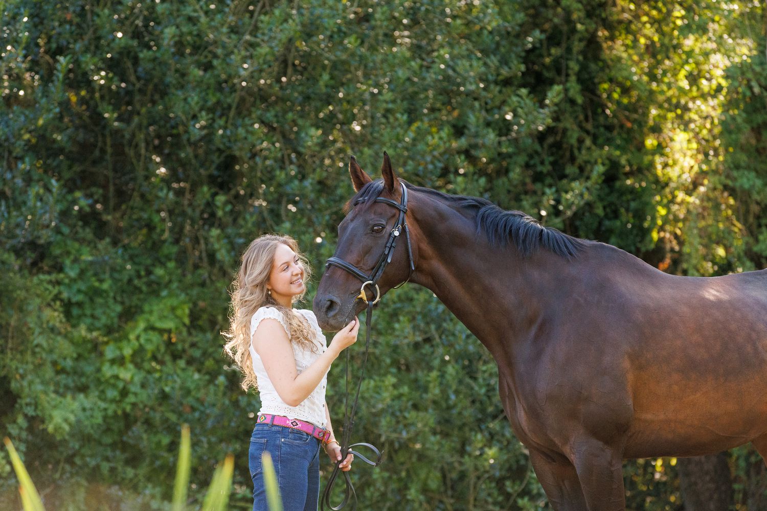 Young woman in black top smiling while gently embracing a brown horse outdoors among lush green trees.