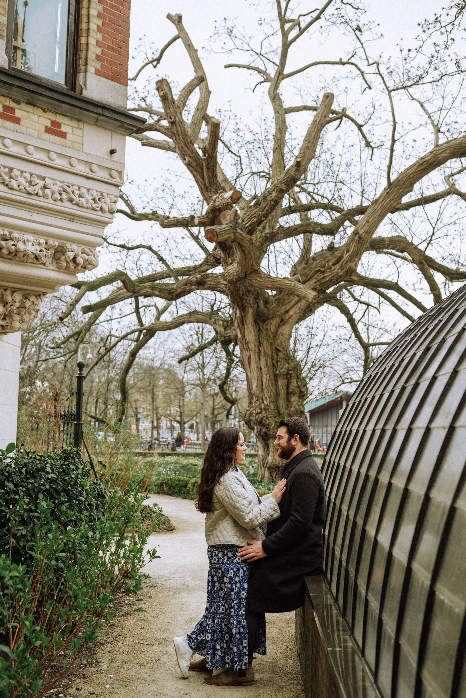 Couple embracing on a garden path beside a large bare tree and a curved greenhouse, with ornate stone architecture nearby.