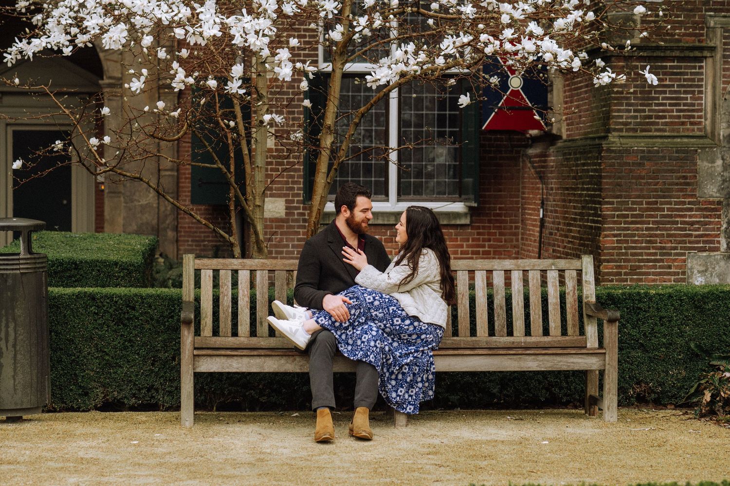Couple sitting close together on a wooden park bench beneath a blooming white magnolia tree in front of a brick building.