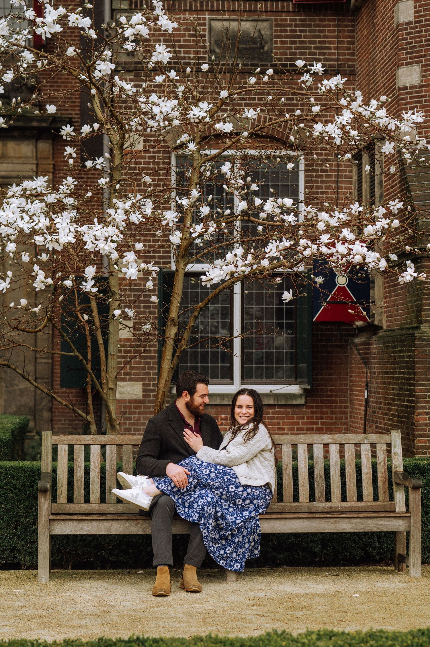 Smiling couple sits close together on a wooden bench beneath a blooming magnolia tree in front of a red brick building.