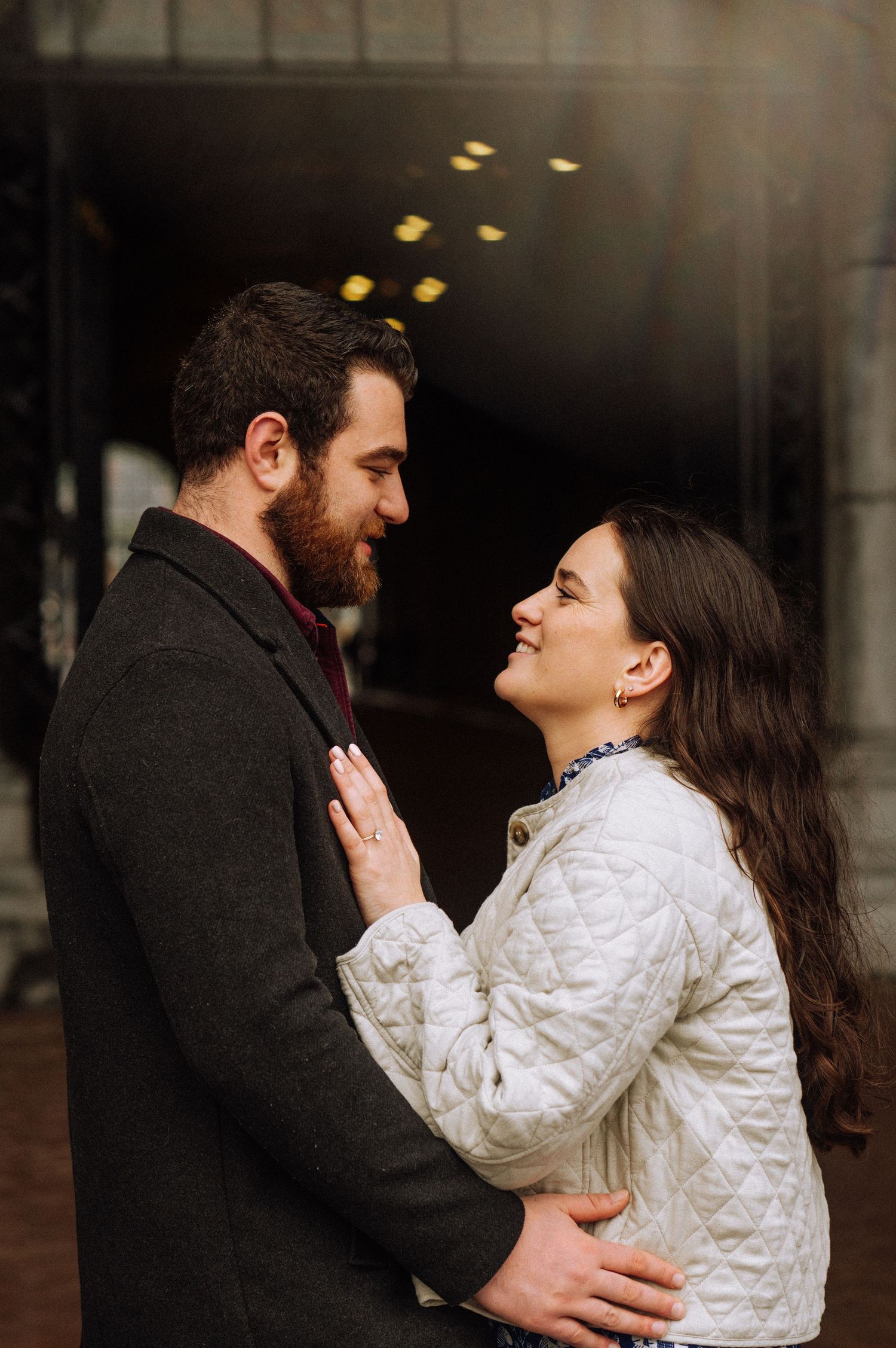 Smiling couple gazing at each other in an engagement photo, the woman in a quilted white jacket showing her ring beside a bearded man.