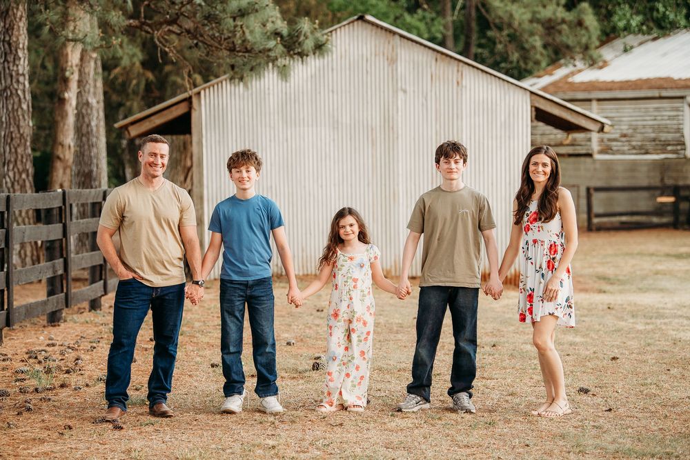 Happy family of five holding hands in front of a rustic barn, posing for an outdoor portrait photo.