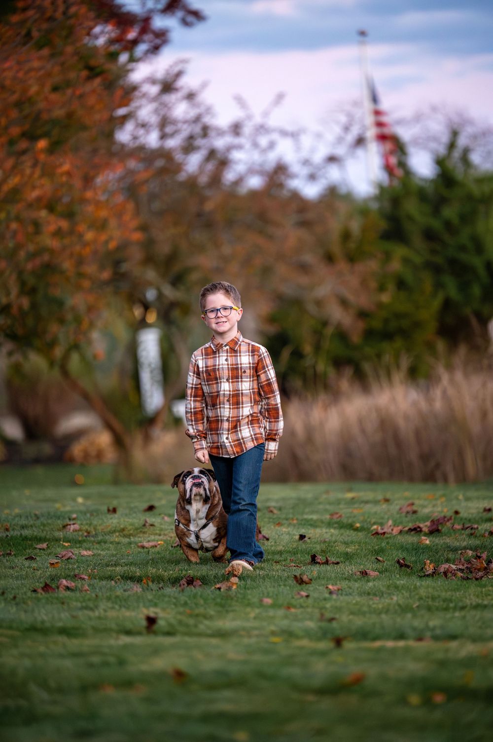 Boy in plaid shirt walks with Basset Hound on green lawn amid fall foliage with American flag in background.