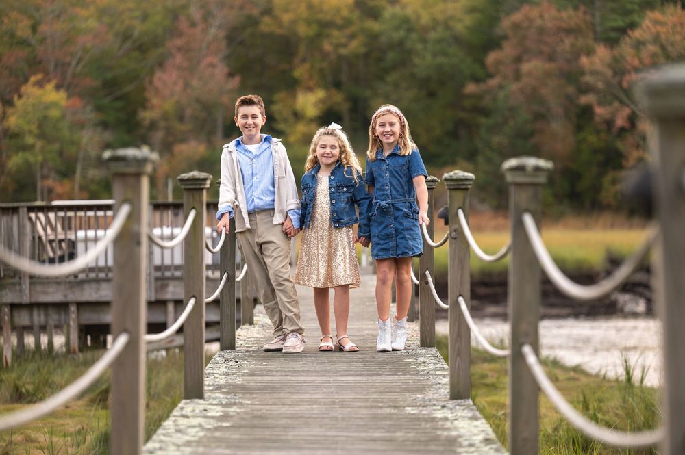 Three children posing on a wooden bridge walkway surrounded by autumn foliage in a family portrait.