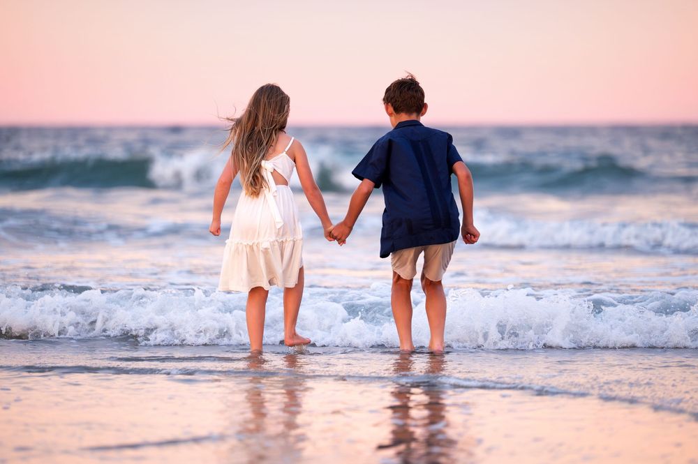 Two children holding hands at the beach during sunset, standing at the water's edge with gentle waves.