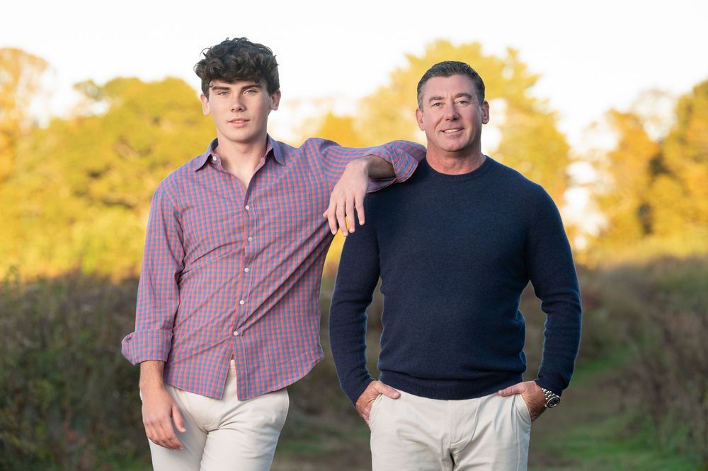 Two men pose outdoors in casual attire against a backdrop of autumn trees in golden light.