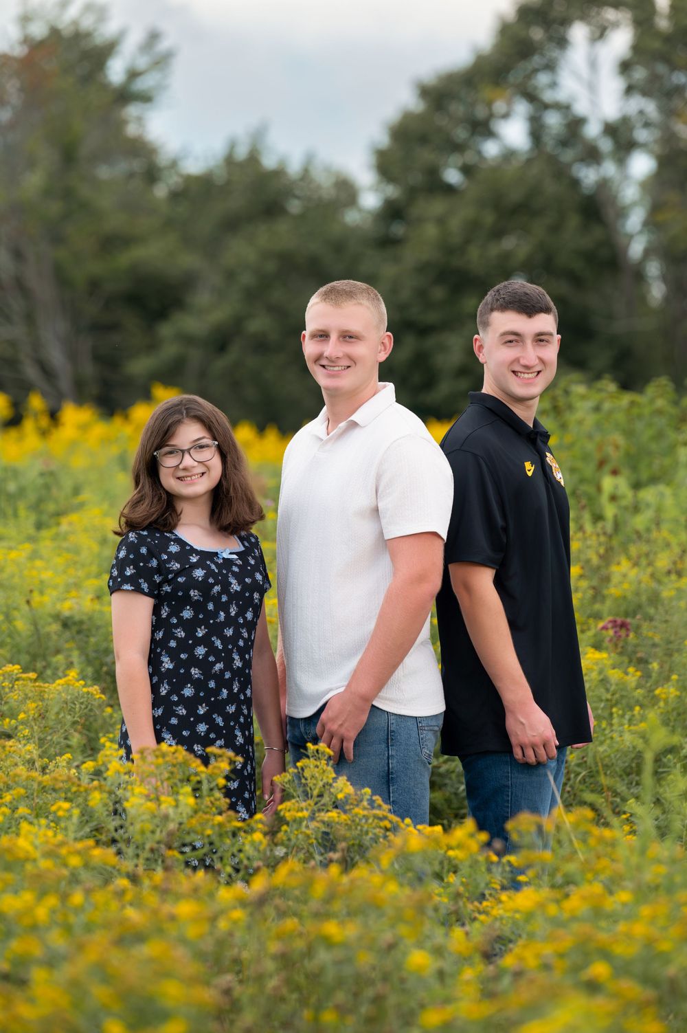 Three young people smiling in a field of yellow wildflowers with trees in the background.