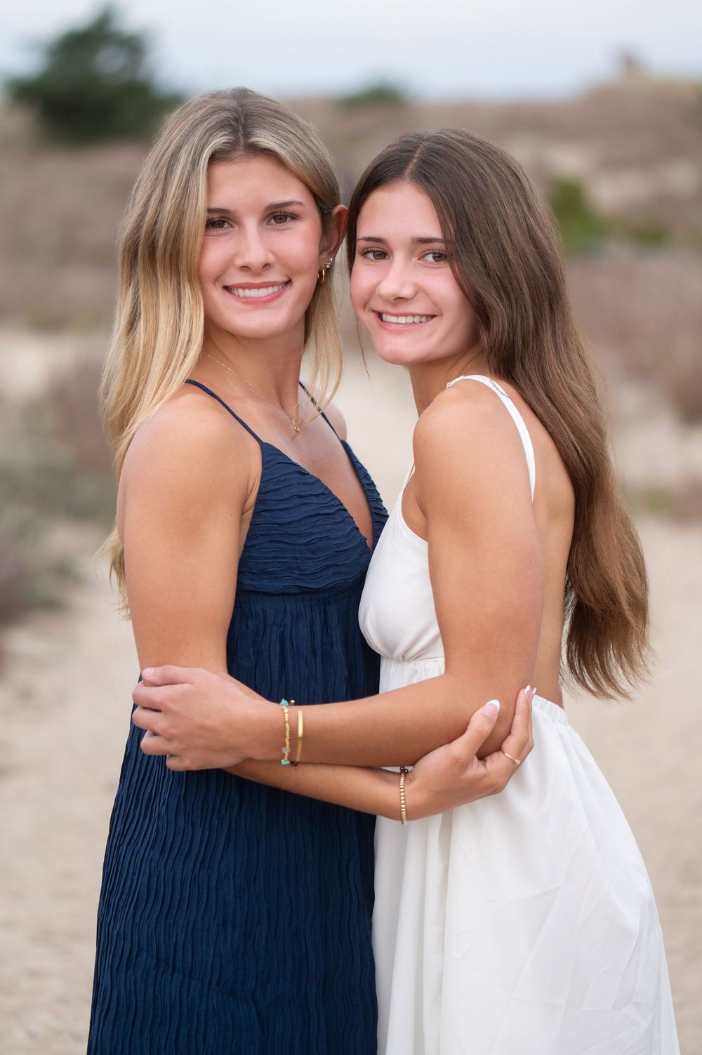 Two smiling young women in navy and white dresses embrace on a sandy beach path.