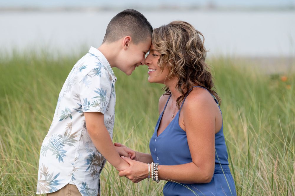 A woman and boy touch foreheads while holding hands in tall grass near water.