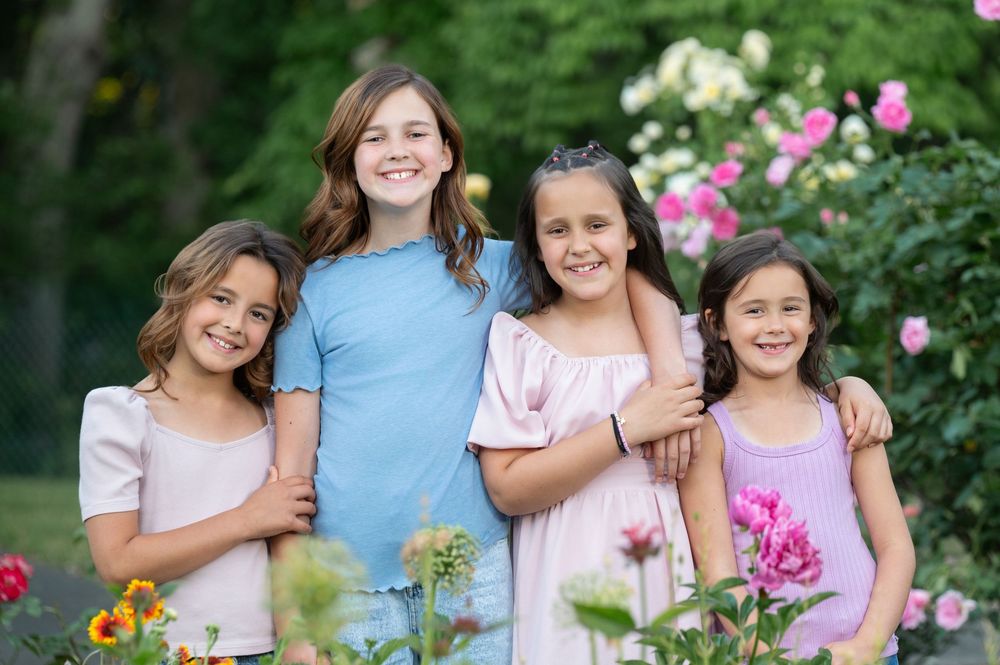 Four smiling girls pose together outdoors among colorful garden flowers and lush green trees.