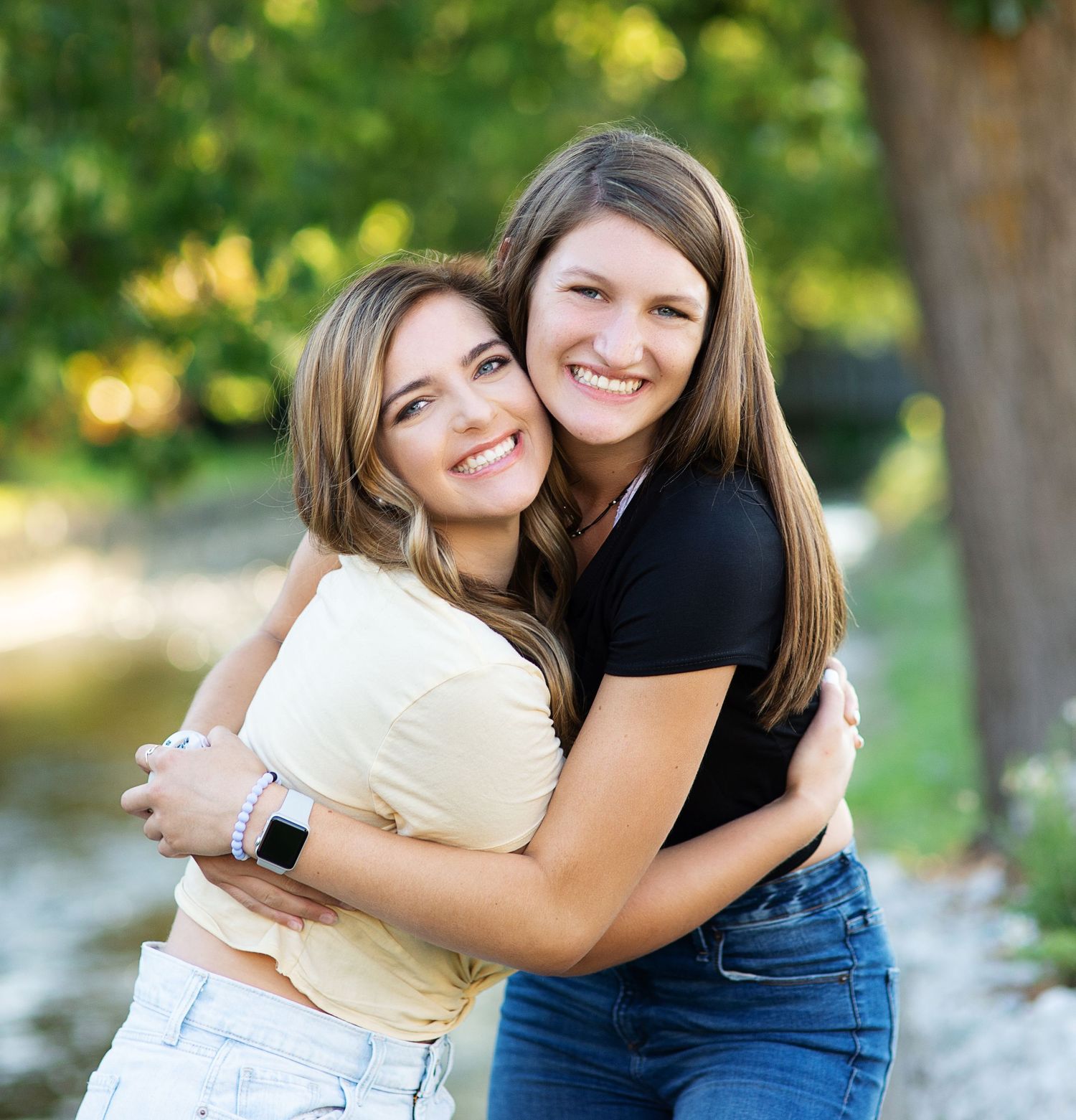 Two teenage girls hugging and smiling outdoors surrounded by green trees.