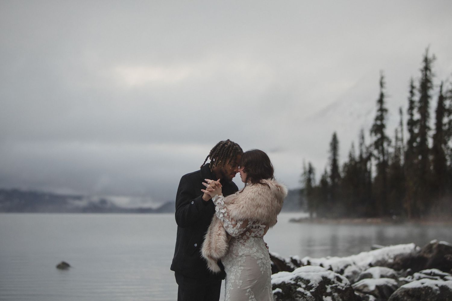 Couple sharing an intimate moment during a snowy winter elopement by a mountain lake surrounded by evergreen trees.