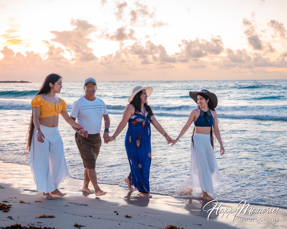 Four people hold hands while walking along a sandy beach at sunset, wearing casual summer outfits and straw hats beside gentle ocean waves.